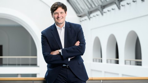 Ein Mann im dunklen Anzug steht im Gang vor einem Lichthof und lächelt in die Kamera.