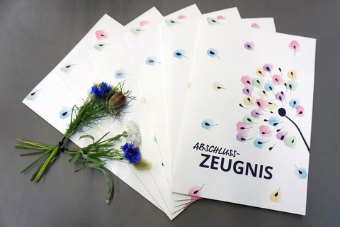 Auf sechs aufgefächerten Zeugnismappen für die Absolventinnen der Universitätsschule Dresden liegt ein Wiesenblumenstrauß.