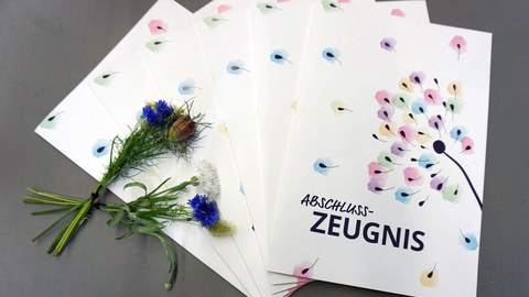 Auf sechs aufgefächerten Zeugnismappen für die Absolventinnen der Universitätsschule Dresden liegt ein Wiesenblumenstrauß.