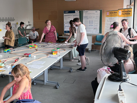 Uniraum. Personen stehen um einen großen Tisch mit aufgebauter Kindereisenbahn im Gespräch, im Hintergrund eine Posterausstellung