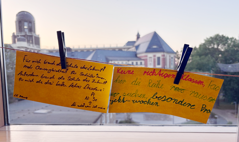 Zwei gelbe Zettel auf einer Wäscheleine mit handschriftlichen Antworten auf die Frage "Wie sieht für dich die Schule der Zukunft aus?"