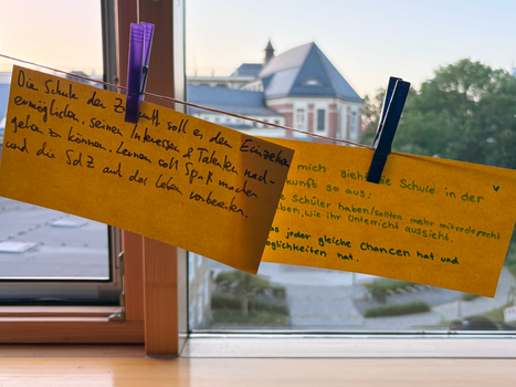 Zwei gelbe Zettel auf einer Wäscheleine mit handschriftlichen Antworten auf die Frage "Wie sieht für dich die Schule der Zukunft aus?"