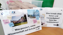Vor einer Kiste mit Holzschienen für eine Kindereisenbahn stehen zwei Schilder mit Logos und Fotos der TU Dresden und der Universitätsschule Dresden und dem Text "Was passiert, wenn Wissenschaft auf Schule trifft?" und "Eine Schule ohne Noten und Hausaufgaben? Kann das funktionieren?"