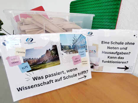 Vor einer Kiste mit Holzschienen für eine Kindereisenbahn stehen zwei Schilder mit Logos und Fotos der TU Dresden und der Universitätsschule Dresden und dem Text "Was passiert, wenn Wissenschaft auf Schule trifft?" und "Eine Schule ohne Noten und Hausaufgaben? Kann das funktionieren?"
