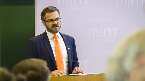 Ein Mann im dunklen Ankzug mit weißem Hemd und orangefarbener Krawatte spricht in einem Hörsaal zum Publikum.