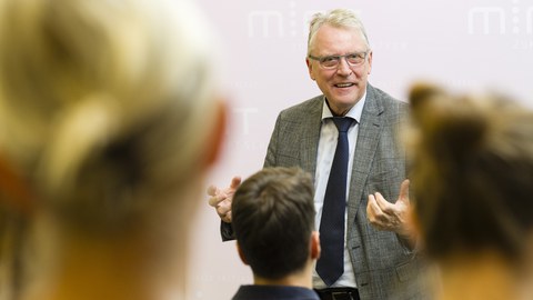 Ein älterer Mann im grauen Anzug spricht begeistert zu einem Publikum im Hörsaal.