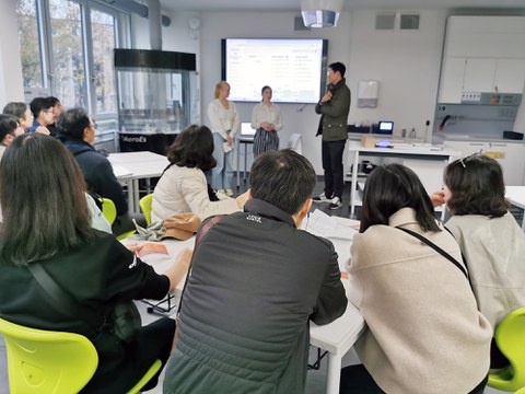 In einemLernraum stehen 2 Schülerinnen und ein Übersetzer vor einer koreanischen Delegation, im Hintergrund ein großer Bildschirm mit individuellem Stundenplan 