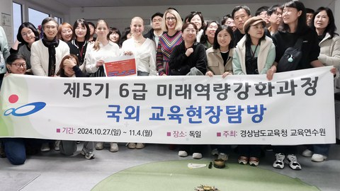 Two pupils and principal Maxi Heß stand surrounded by around 20 people from South Korea behind a banner reading ‘Improve future skills of Grades 5 and 6 - Educational trip through Germany - 27 Oct - 4 Nov 2024 - Gyeongsangnam-do Education Office