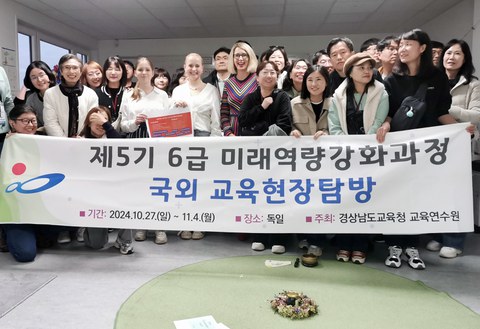 Zwei Schülerinnen und Schulleiterin Maxi Heß stehen umringt von etwa 20 Personen aus Südkorea hinter einem Banner mit der Aufschrift " Verbesserung zukünftiger Fähigkeiten der Klassen 5 und 6 – Bildungsreise durch Deutschland - 27.10.-4.11.2024" 