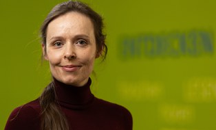 Portrait einer Mitarbeiterin der Forschungsstelle ForUs zur Universitätsschule Dresden. Eine brünette Frau mit auberginefarbenem Rollkragenpullover schaut in die Kamera. Im Hintergrund sind auf einer grünen Wand die Wörter Entdecken, Lesen, Hören, Rechnen verschwommen zu erkennen.