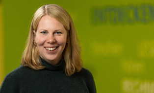 Portrait einer Mitarbeiterin der Forschungsstelle ForUs zur Universitätsschule Dresden. Eine brünette Frau mit auberginefarbenem Rollkragenpullover schaut in die Kamera. Im Hintergrund sind auf einer grünen Wand die Wörter Entdecken, Lesen, Hören, Rechnen verschwommen zu erkennen.