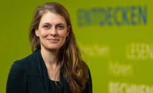 Portrait einer Mitarbeiterin der Forschungsstelle ForUs zur Universitätsschule Dresden. Eine brünette Frau mit auberginefarbenem Rollkragenpullover schaut in die Kamera. Im Hintergrund sind auf einer grünen Wand die Wörter Entdecken, Lesen, Hören, Rechnen verschwommen zu erkennen.