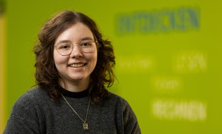 Portrait einer Mitarbeiterin der Forschungsstelle ForUs zur Universitätsschule Dresden. Eine brünette Frau mit auberginefarbenem Rollkragenpullover schaut in die Kamera. Im Hintergrund sind auf einer grünen Wand die Wörter Entdecken, Lesen, Hören, Rechnen verschwommen zu erkennen.