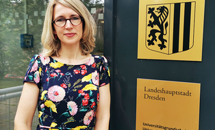 Maxi Heß steht vor der Schuleingangstür, neben ihr zwei goldene Schilder mit dem Wappen der Stadt Dresden und der Aufschrift Universitätsgrundschule und Universitätsoberschule.