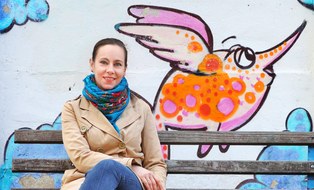 Auf einer Bank vor einer bunten Grafittiwand an der Universitätsschule Dresden sitztKommunikationsamangerin Maria Völzer.