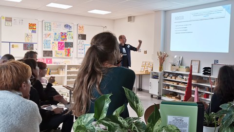 Eine Workshop-Gruppe schaut auf einen Lernbegleiter. Er erklärt ein an die Wand projiziertes Schaubild.