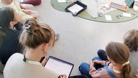 Vogelperspektive auf sitzende Kinder im Kreis. Eine junge Frau sitzt mit im Kreis macht auf dem Tablet Notizen. In der Mitte ein grüner runder Teppich mit Lernmaterialien.