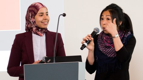 Youmna Fouad und Mei-Chen Spiegelberg tragen vor, stehen hinter einem Pult, Mei-Chen spricht in ein Mikrofon
