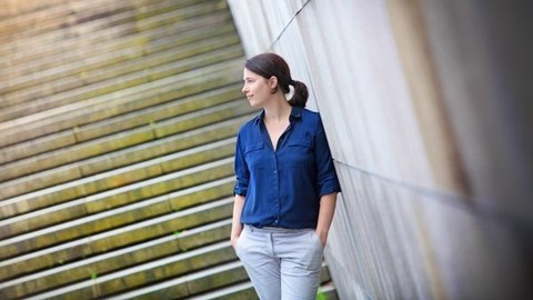 Foto von Sabine Müller-Mall, stehend vor einer Treppe angelehnt an eine Mauer, mit Händen in den Hosentaschen, nach links blickend