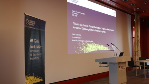 Leere Bühne mit Stehpult und Beamerpräsentation des Vortrags von Prof. Dr. Heike Greschke und Youmna Fouad