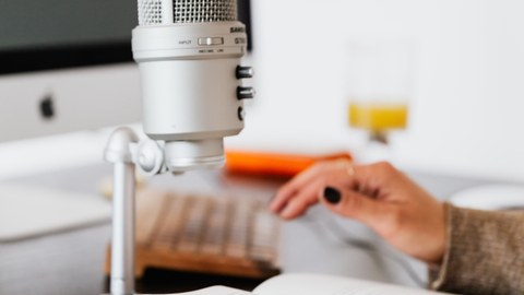 Farbfoto eines Mikrofons, welches auf einem Tisch steht. Auf dem Tisch sind ein Notizheft sowie der Auschnitt zweier Hände zu sehen. 