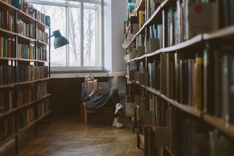 Foto einer jungen Frau in einer Bibliothek. Sie liegt entspannt in einem Stuhl und liest. Die Regale links und rechts im Bild sind leicht verschwommen.