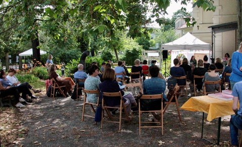 Im Garten ist ein Pavillion aufgebaut. Jemand liest etwas vor. Menschen sitzen auf Stühlen um den Pavillion herum. Es scheint die Sonne.