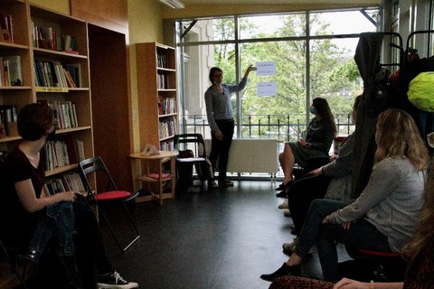 In a room with many bookshelves, people sit on chairs and listen to a woman standing at the window showing something.