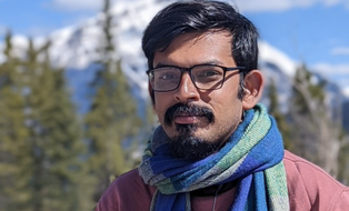Portrait of a man with beard, glasses, and a blue scarf with a mountain landscape in the background