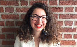 Portrait of a woman with shoulder-length, brown, curly hair, glasses, and earrings wearing a white shirt standing in front of a brick wall.