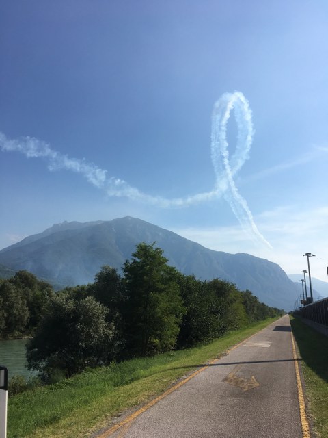 Fahrradweg mit Blick auf einen Berg. Darüber hat ein Flieger ein Looping in den Himmel gezeichnet.
