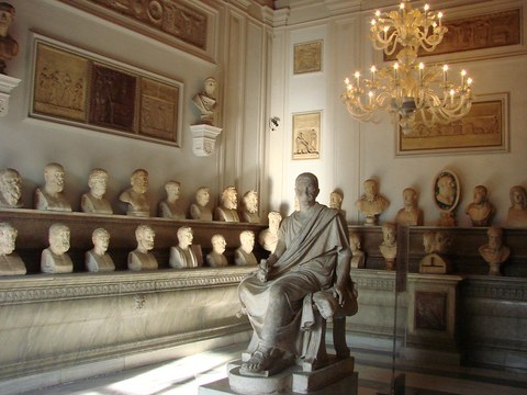 Saal in klassischer Architektur mit Büsten auf zwei umlaufenden Regalen, in der Mitte die Skulptur eines sitzenden Konsuls.