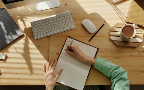 Jemand notiert etwas in ein Notizbuch, das auf einem Schreibtisch liegt. Rechts steht eine Tasse Milchkaffee. Die Sonne scheint auf den Schreibtisch. Eine Apple-Tastatur liegt vor dem Notizbuch.