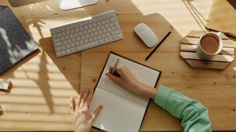 Jemand notiert etwas in ein Notizbuch, das auf einem Schreibtisch liegt. Rechts steht eine Tasse Milchkaffee. Die Sonne scheint auf den Schreibtisch. Eine Apple-Tastatur liegt vor dem Notizbuch.