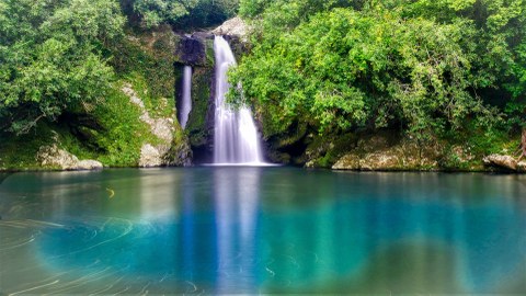 Zu sehen ist ein intensiv türkis-blaues Wasserbecken, in welches ein Wasserfall mündet. Umgeben ist die Szenerie von tropischen Pflanzen in leuchtend-sattem Grün.