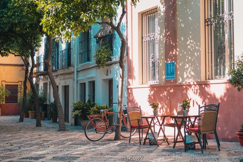 Ein sonniger Platz in Südeuropa mit einer Sitzgruppe und einem Fahrrad, dass an einer bunten Hauswand lehnt. 