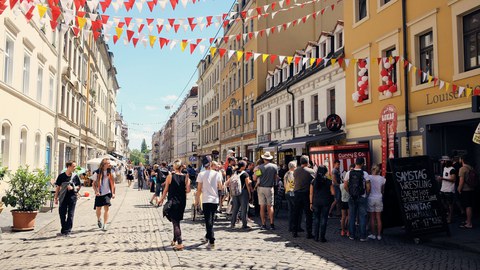 Zu sehen ist eine helle Straße, auf der Menschen laufen und stehen. Zwischen den Häusern links und rechts sind bunte Wipfel gespannt.