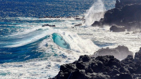 Zu sehen sind Wellen, welche sich an vor der Küste liegenden Felsen brechen.