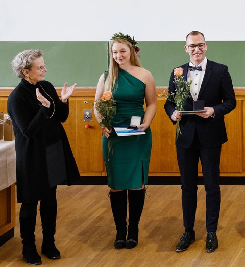 Links im Bild steht die Rektorin Prof. Dr. Staudinger und applaudiert den ausgezeichneten Studierenden. Neben ihr steht Rose Sharon Király in einem grünen Kleid und einer Blume in der Hand, rechts neben ihr steht ein weitere Student im Anzug.