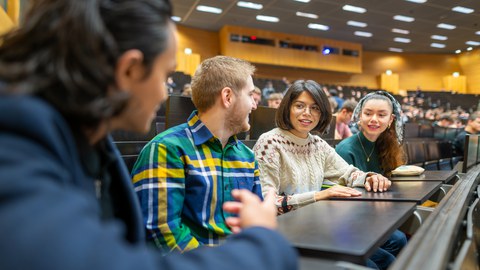 Fotos von vier sich unterhaltenden Studierenden in einem vollen TUD-Hörsaal.