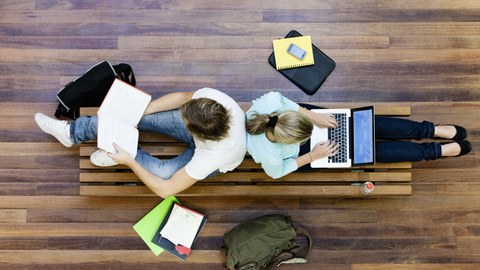 Zwei Studierende sitzen mit Büchern und Laptop am Fußboden