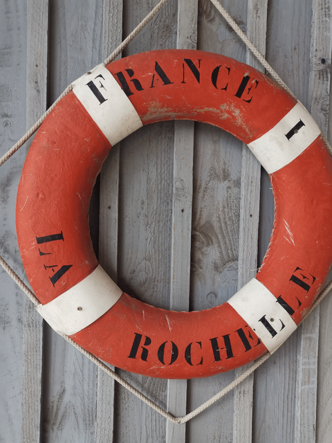 Das Foto stellt einen roten Schwimmring dar, welcher an einer grauen Holzwand aufgehängt ist; unterbrochen von vier senkrechten weißen Streifen. Oben der Schriftzug "FRANCE" und unten "LA ROCHELLE".