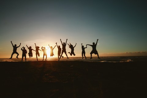Elf Personen, die vorm Sonnenuntergang am Strand in die Luft springen. Im Hintergrund sieht man das Meer.