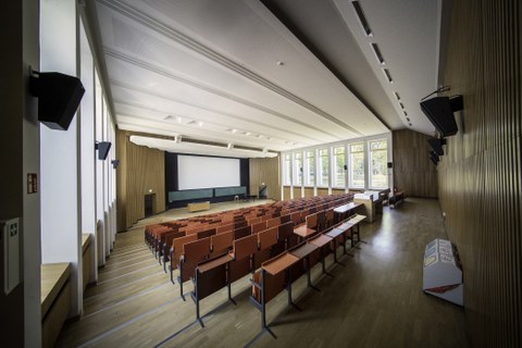 Leerer Hörsaal in der August-Bebel-Straße. Im Vordergrund befindet sich die Stuhlreihen, im Hintergrund das Rednerpult