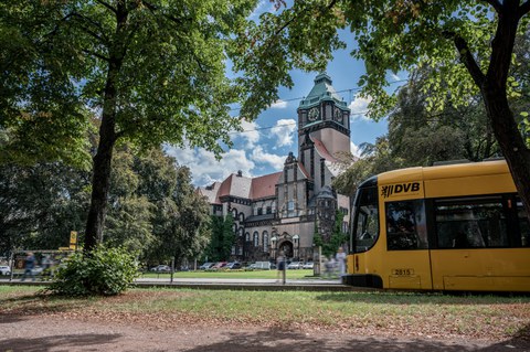 Foto vom Georg-Schumann-Bau und der Haltestelle davor mit einer Straßenbahn und den Fußgängern