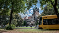 Foto vom Georg-Schumann-Bau und der Haltestelle davor mit einer Straßenbahn und den Fußgängern