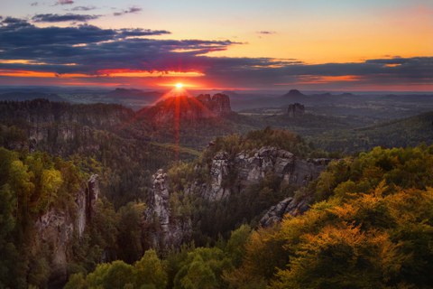 Alternativtext Das Foto ist eine Panoramaaufnahme der Sächsischen Schweiz bei Dämmerung. Die Landschaft ist geprägt von grün bewachsenen hohen Felsen die sich bis in die Ferne ziehen. Das obere Drittel des Fotos ist von einem bewölkten rot-orangen Himmel bedeckt, aus dem die rote Sonne aus einem Wolkenschlitz scheint.