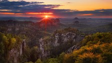 Alternativtext Das Foto ist eine Panoramaaufnahme der Sächsischen Schweiz bei Dämmerung. Die Landschaft ist geprägt von grün bewachsenen hohen Felsen die sich bis in die Ferne ziehen. Das obere Drittel des Fotos ist von einem bewölkten rot-orangen Himmel bedeckt, aus dem die rote Sonne aus einem Wolkenschlitz scheint.