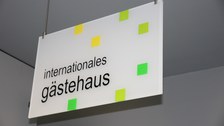 Weißes Schild mit kleinen gelben und grünen Quadraten. Darauf befindet sich die Aufschrift: "internationales gästehaus"