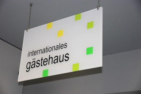 Weißes Schild mit kleinen gelben und grünen Quadraten. Darauf befindet sich die Aufschrift: "internationales gästehaus"